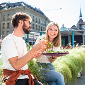 Mann (Links) und Frau (Rechts) sitzen zwische Pflanzen und geniessen ihre Pizza aus der reCIRCLE Pizza BOX